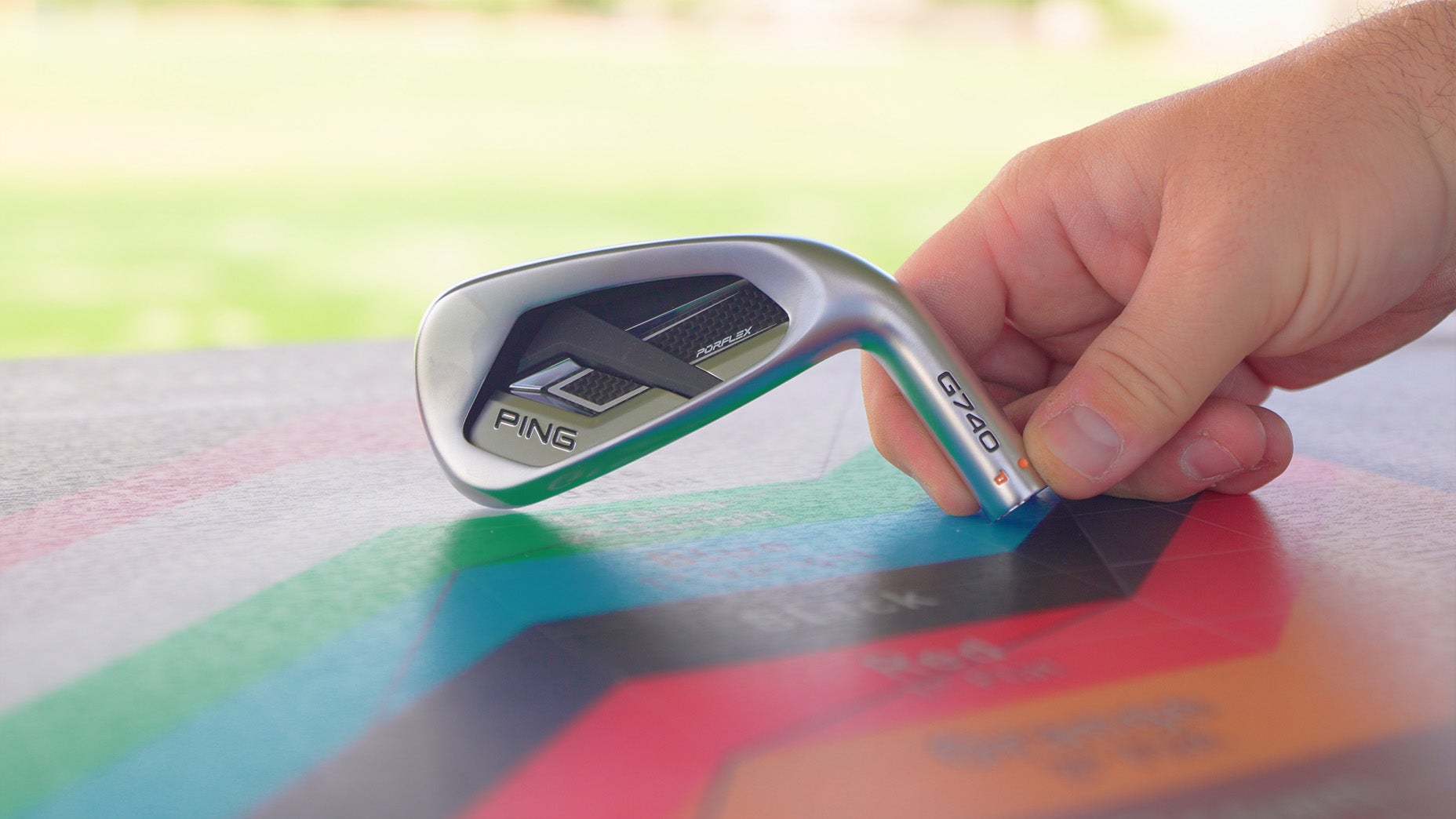 A hand holding a PING G740 golf iron resting on a colorful, hexagon-patterned surface with a blurred grassy background.