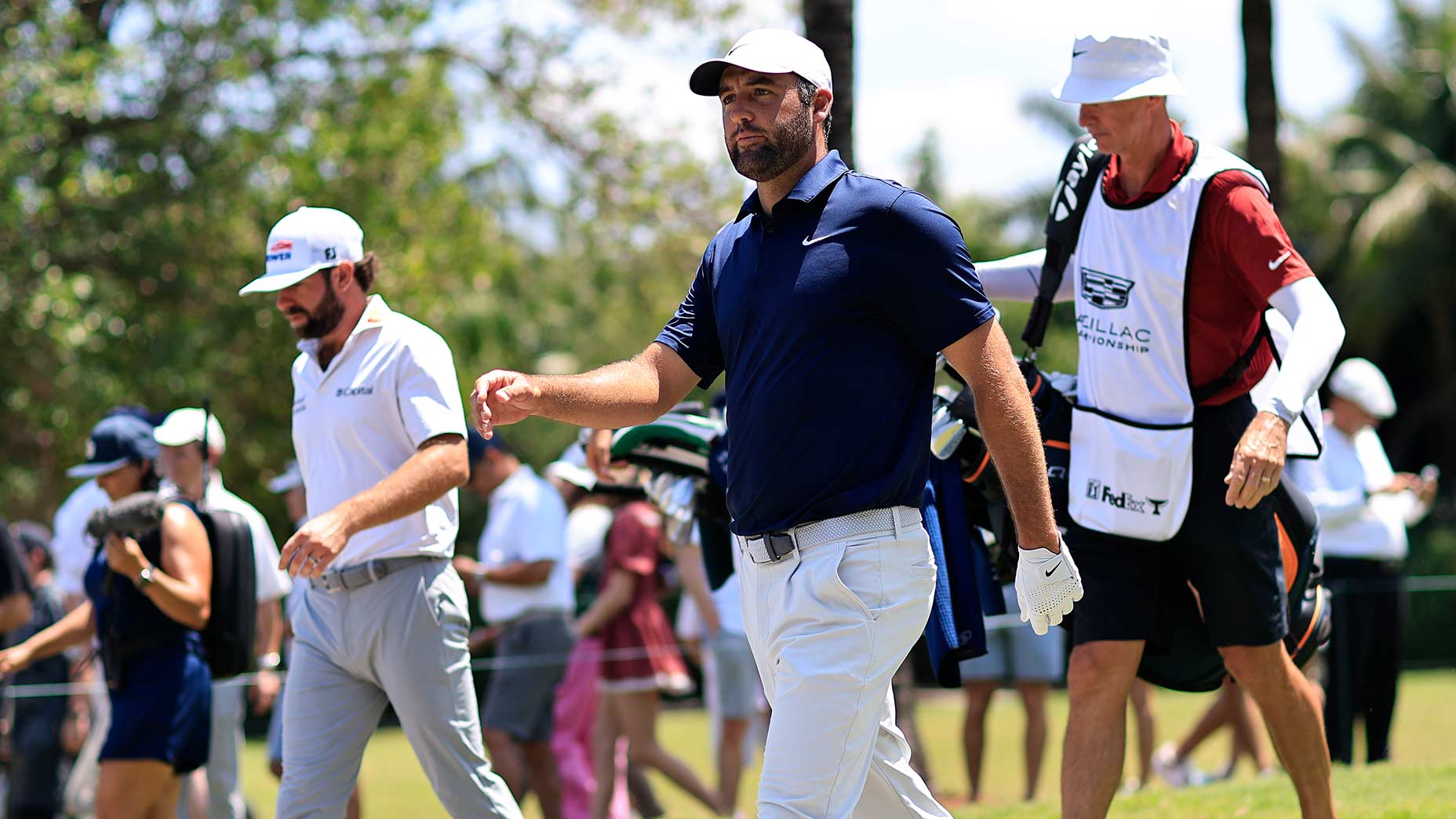 Two golfers walk on a sunny LIV Golf course, each holding clubs, with a caddie in a white bib following behind. Spectators and trees are visible in the background as players react to their shots.