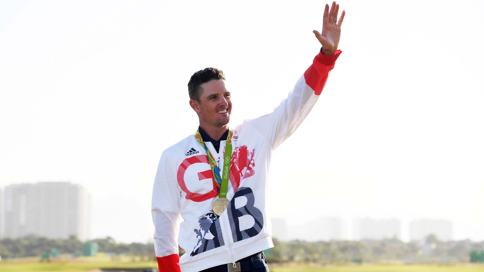 A male athlete, Justin Rose, wearing a Team GB jacket and three medals around his neck smiles and waves with his right hand. He stands outdoors with city buildings in the background.