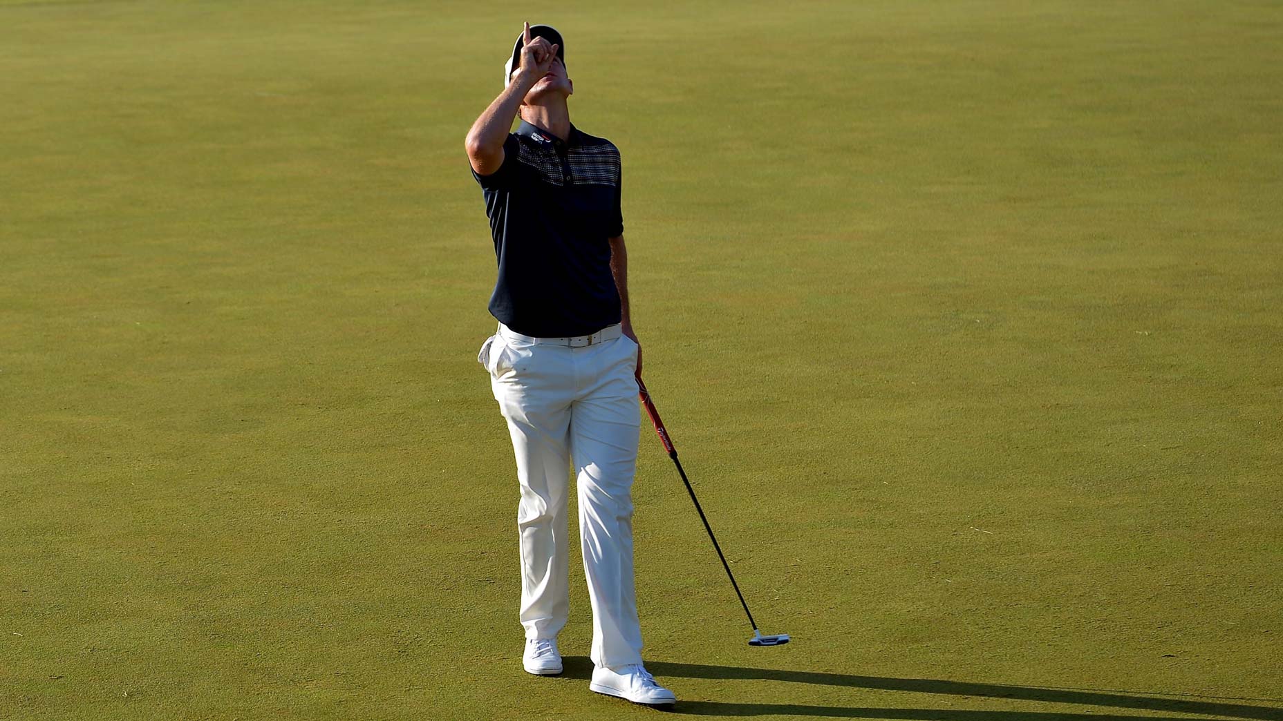 A golfer wearing a black shirt and white pants stands on a green golf course, holding a putter and pointing upward like Justin Rose after a shocking move, while looking at the sky.